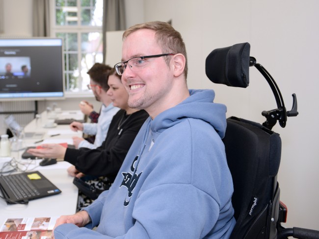 Ein Mann im Rollstuhl sitzt an einem Schreibtisch und lächelt, er und andere Teilnehmer nehmen an einer Videokonferenz teil